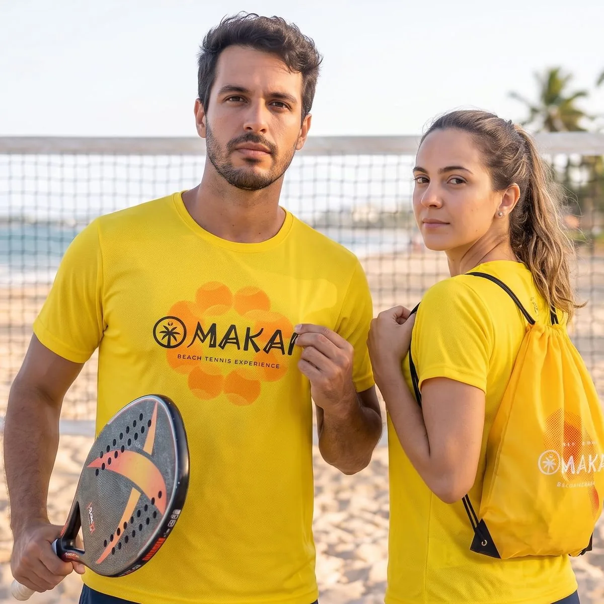 Dupla de atletas na praia, em frente à rede, vestindo a camiseta oficial amarela do Makai; ela leva a sacochila amarela nas costas